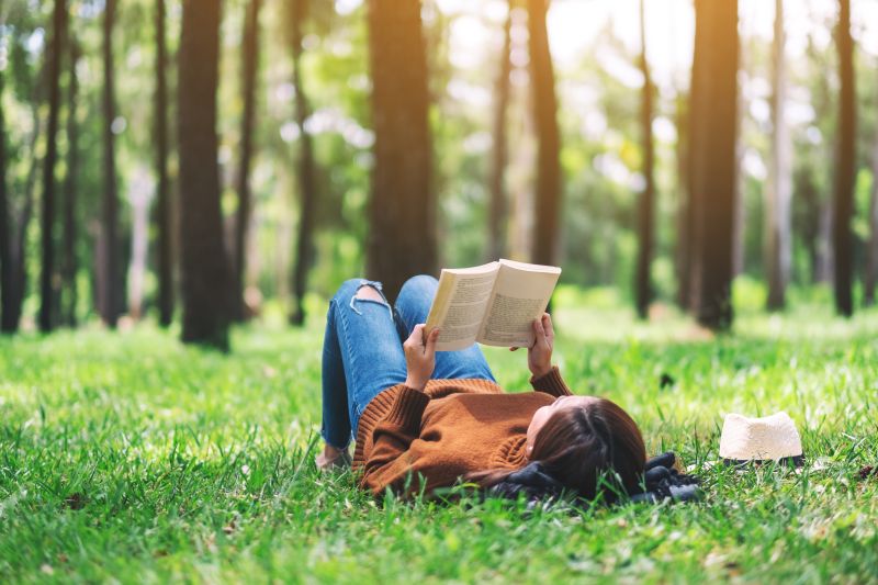 Person lying on grass, reading a book. There are trees in the backgroun.